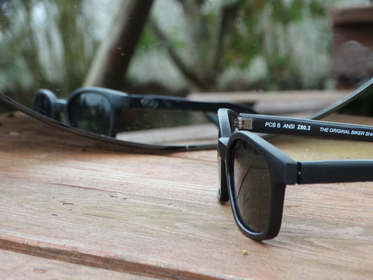 Sunglasses KD's smoked lenses, matte black frame posed in front of a mirror.
