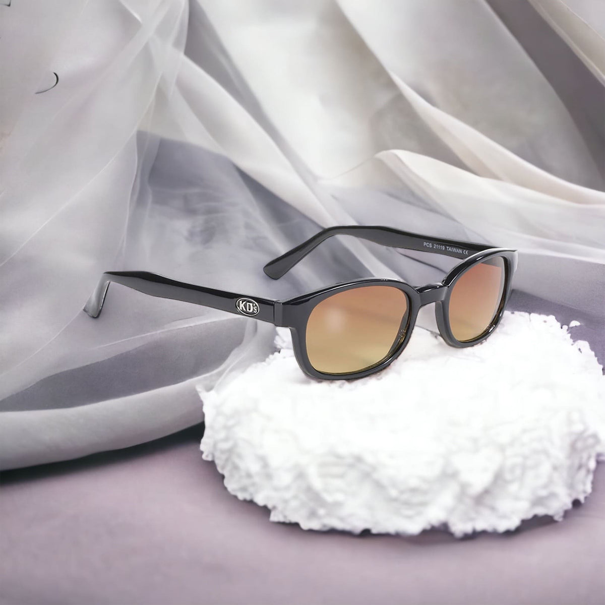 KD's 21119 sunglasses that fit under a motorcycle or ski helmet. For outdoor activities. With a stylish black frame and amber lenses. Placed in front of curtains.