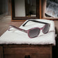 The KD's 2116 sunglasses perfect for sunny days with grey lenses and an elegant pink frame laid on a soft blanket in a wooden house.