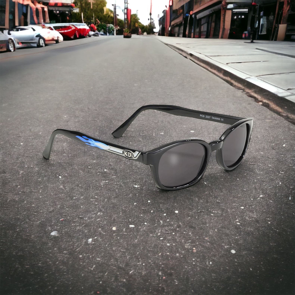 The KD's 2227 sunglasses with grey polycarbonate lenses and a frame with a blue flame-emitting tailpipe pattern placed in the street in front of cars.