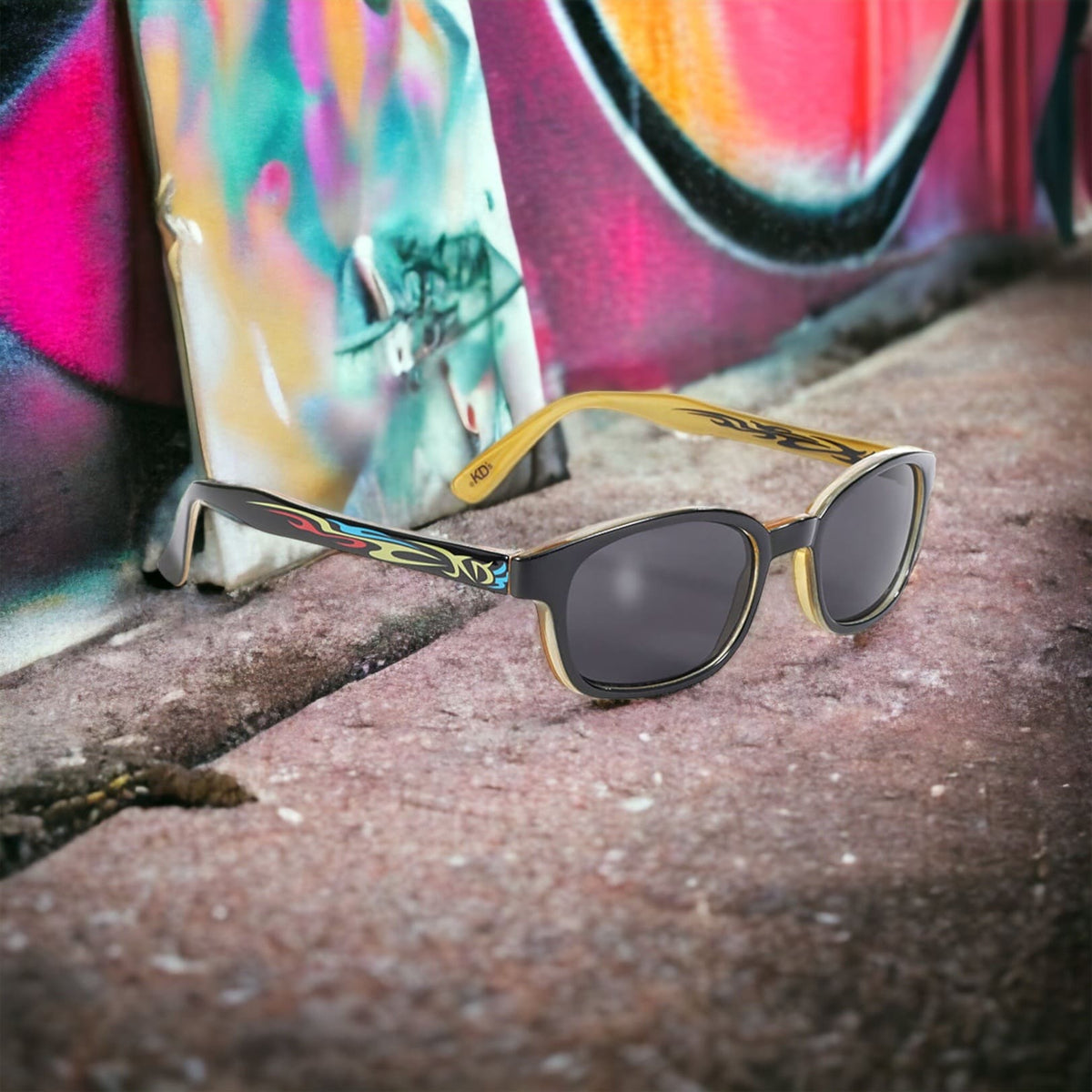 The KD's 5400 sunglasses with grey polycarbonate lenses and a black frame with a light blue and red tribal tattoo pattern exposed on a sidewalk behind graffiti.